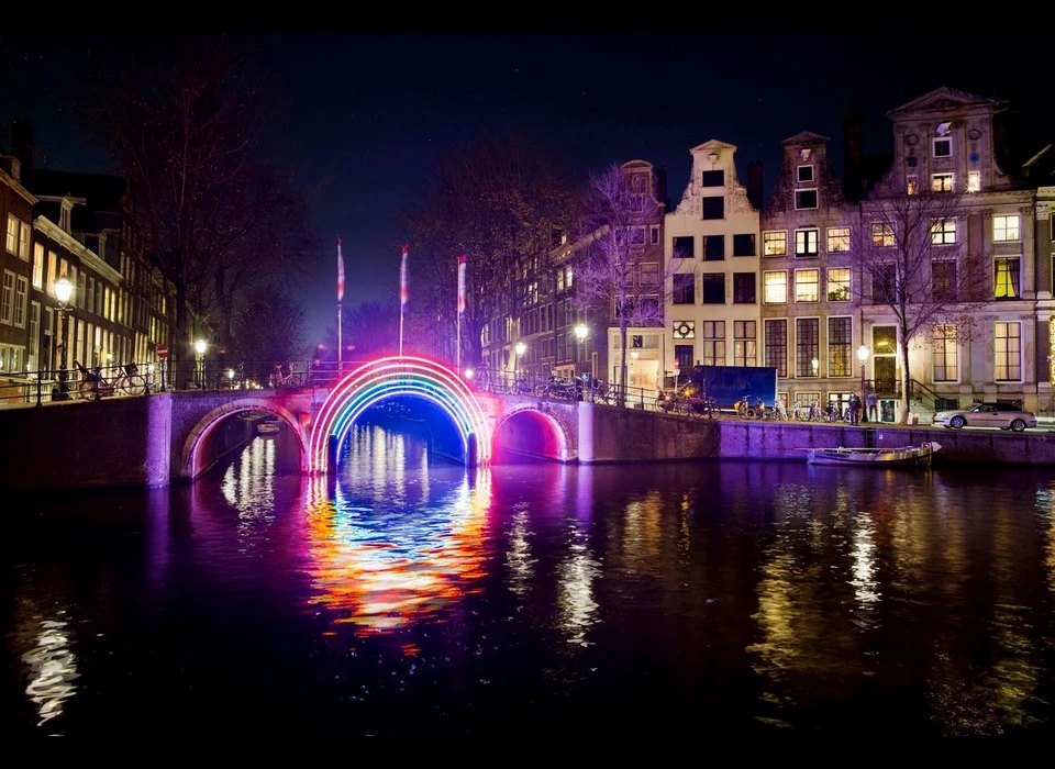 De filosoof René Descartes (1596-1650) wist, toen hij in 1636 in Amsterdam woonde, als eerste een regenboog wetenschappelijk te verklaren. 
					  Een regenboog is normaliter ongrijpbaar maar Gilbert Moity zijn regenboog is echter wél tastbaar en contrasteert met de donkere nacht. Hij creëert met het werk een verbinding tussen twee oevers en een poort. Het werk 
					  symboliseert zo de openheid en verscheidenheid die typisch zijn voor Amsterdam. Het kleurenschema staat ook voor LGBTQIA (Lesbian, Gay, Bisexual, Transgender, Queer, Intersex, Asexual), een belangrijke community 
					  in de stad. (Gilbert Moity (FR) werkt graag samen met ambachtslieden. Hij gebruikt licht als belangrijkste instrument van zijn artistieke expressie. Elke keer wanneer hij aan een nieuw project begint, probeert Moity de 
					  grenzen van technologie te verleggen.) 2016