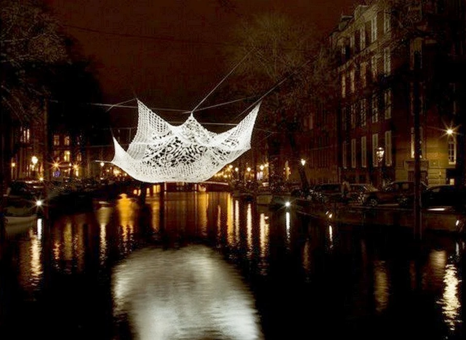 'The lace' zou een vliegend tapijt kunnen zijn: het geeft een zweverig en magisch gevoel als je ernaar kijkt. In werkelijkheid is het een enorm 
					  stuk kant (in het Engels ‘lace’) dat een traditioneel Noord-Hollands kapje symboliseert. Het werk is gemaakt van draden die licht reflecteren. Het symboliseert de dicht verweven stad: de verwevenheid van verschillende 
					  mensen, de stedelijke structuur en de symbiose van historisch Amsterdam met de multiculturele stad van vandaag. Choi verwerkte aspecten van haar Koreaanse achtergrond in het werk (Hanbok, een traditionele naai- en 
					  vouwpapierkunst), waarmee ze de waarde van culturele kruisbestuiving toont. (Choi + Shine Architects (VS) is een internationale designstudio, opgericht door Jin Choi en Thomas Shine. Choi studeerde Architectural Art 
					  aan Yale University. Ze werd in 2013 uitgeroepen tot ‘Global Young Architect’. Thomas Shine was als docent Architectural Structures aan Yale University verbonden.) 2016