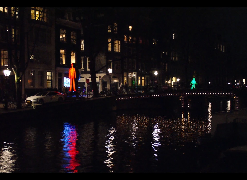 De mannetjes uit het alledaagse voetgangerslicht (een rood, een groen), zijn in de installatie Strangers in the Light meer dan pictogrammen. De mannetjes worden, door hun formaat van 6 meter hoog, personages in een eigen verhaal: 
					  tot leven gekomen en ontsnapt uit hun ‘zwarte doos’ ontmoeten ze elkaar op de Herengracht voor het eerst. Want wanneer er eentje verscheen, verdween de ander, en zo ontglipten ze elkaar gevangen in het stoplicht 
					  steeds weer. Wie weet raken ze nu eindelijk bevriend of kan er zelfs een romance ontstaan, zoals Frank Sinatra het bezong in het liedje dat de titel van dit werk inspireerde: We were strangers in the night / Up to 
					  the moment when we said our first hello / Little did we know / Love was just a glance away, a warm embracing dance away. (Victor Engbers (NL) schrijft teksten, maakt sculpturen, grafisch werk en treedt op als performancekunstenaar. 
					  Het grootste deel van zijn werk speelt zich af rondom het thema: de wereld niet voor lief nemen. Ina Smits (NL) is beeldend kunstenaar.) 2018