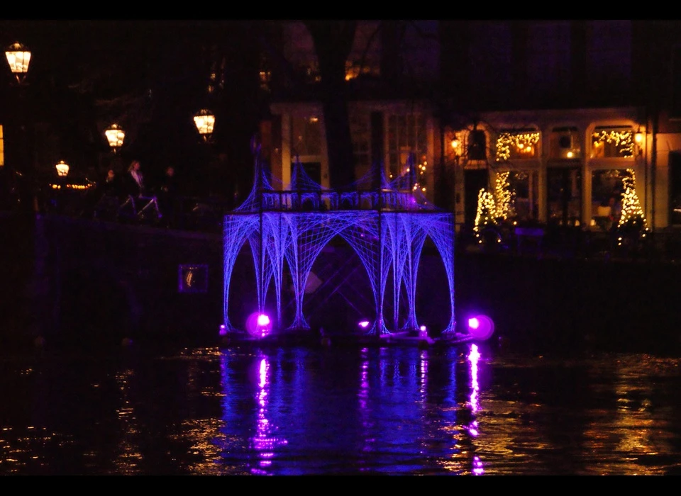 Een spookachtig, blauw-paars verlicht paviljoen ligt in het water aan het einde van de Herengracht, als het restant van een verloren, verdronken kathedraal: het is Peter Snijders installatie 'ArchesTextures: Portam Civitatis'.
					  Met zijn puntige torentjes en vele bogen herinnert deze stadspoort je misschien wel aan de gotische kerken die tussen 1140 en 1500 overal in Europa werden gebouwd. Denk hierbij aan de Oude Kerk in Amsterdam. 'Portam Civitatis' bestaat 
					  uit een simpele constructie, bekleed met fragiele draden die als getekende lijnen oplichten door uv-lampen. (De Belgische Peter Snijder (1974) is een autodidactische ‘kunstenaar-uitvinder’, zoals hij zichzelf omschrijft, 
					  die altijd licht verwerkt in zijn kunstwerken. Hij gebruikt het liefst gevonden of gekregen materialen en bouwt zijn architectuur-achtige constructies, ontworpen in zijn persoonlijke, fantasierijke vormentaal, zelf.) 2018