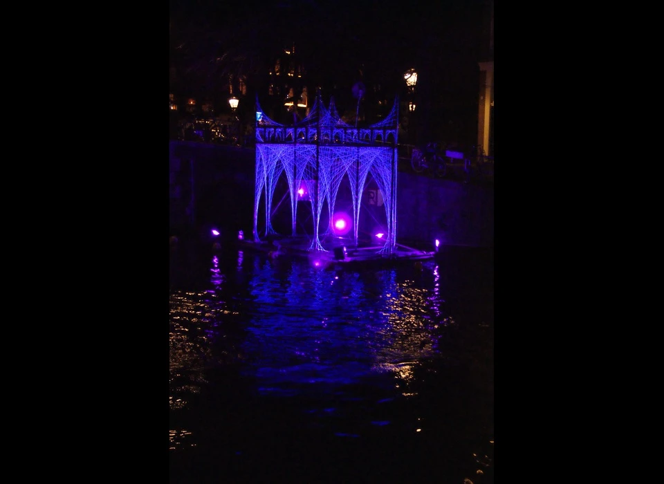 Een spookachtig, blauw-paars verlicht paviljoen ligt in het water aan het einde van de Herengracht, als het restant van een verloren, verdronken kathedraal: het is Peter Snijders installatie 'ArchesTextures: Portam Civitatis'.
					  Met zijn puntige torentjes en vele bogen herinnert deze stadspoort je misschien wel aan de gotische kerken die tussen 1140 en 1500 overal in Europa werden gebouwd. Denk hierbij aan de Oude Kerk in Amsterdam. 'Portam Civitatis' bestaat 
					  uit een simpele constructie, bekleed met fragiele draden die als getekende lijnen oplichten door uv-lampen. (De Belgische Peter Snijder (1974) is een autodidactische ‘kunstenaar-uitvinder’, zoals hij zichzelf omschrijft, 
					  die altijd licht verwerkt in zijn kunstwerken. Hij gebruikt het liefst gevonden of gekregen materialen en bouwt zijn architectuur-achtige constructies, ontworpen in zijn persoonlijke, fantasierijke vormentaal, zelf.) 2018