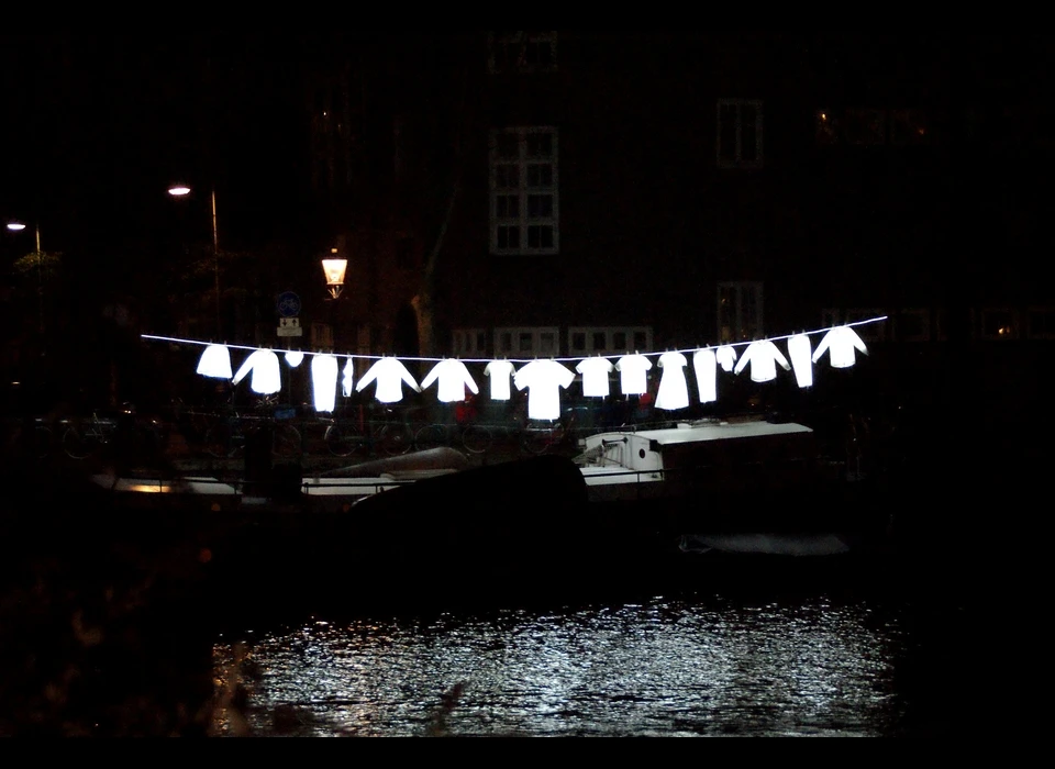 Lichtgevend wasgoed hangt aan waslijnen te wapperen 
					  alsof het een zomerse dag is in Amsterdam. De wit oplichtende kledingstukken, een vrolijke verzameling hemden, T-shirts, ondergoed, 
					  broeken en jurken, hangen aan weerszijde van de gracht. Ook zijn er enkele bijzondere kledingstukken te zien zoals een Turkse 
					  wijde broek, een traditionele Joodse jurk of een Marokkaanse djellaba. Samen vertegenwoordigen deze stukken de culturele en 
					  etnische mix van bewoners van de stad. Verspreid tussen verschillende typisch Amsterdamse woonboten is Neighborhood een subtiele 
					  maar verrassende ingreep in het straatbeeld. Het is een ontwerp van Sergey Kim. 2019