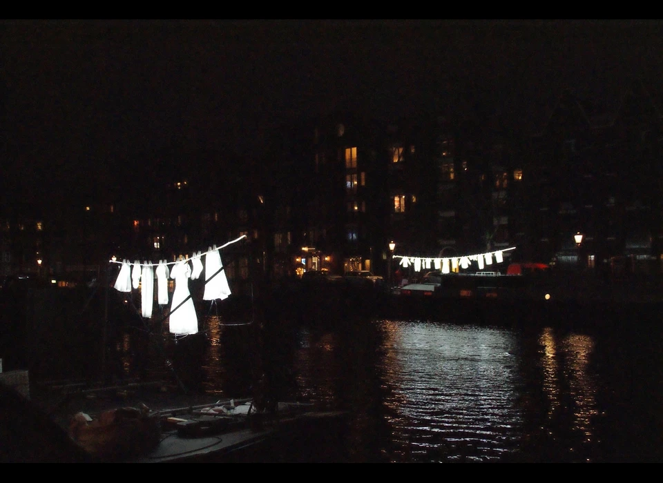 Lichtgevend wasgoed hangt aan waslijnen te wapperen 
					  alsof het een zomerse dag is in Amsterdam. De wit oplichtende kledingstukken, een vrolijke verzameling hemden, T-shirts, ondergoed, 
					  broeken en jurken, hangen aan weerszijde van de gracht. Ook zijn er enkele bijzondere kledingstukken te zien zoals een Turkse 
					  wijde broek, een traditionele Joodse jurk of een Marokkaanse djellaba. Samen vertegenwoordigen deze stukken de culturele en 
					  etnische mix van bewoners van de stad. Verspreid tussen verschillende typisch Amsterdamse woonboten is Neighborhood een subtiele 
					  maar verrassende ingreep in het straatbeeld. Het is een ontwerp van Sergey Kim. 2019