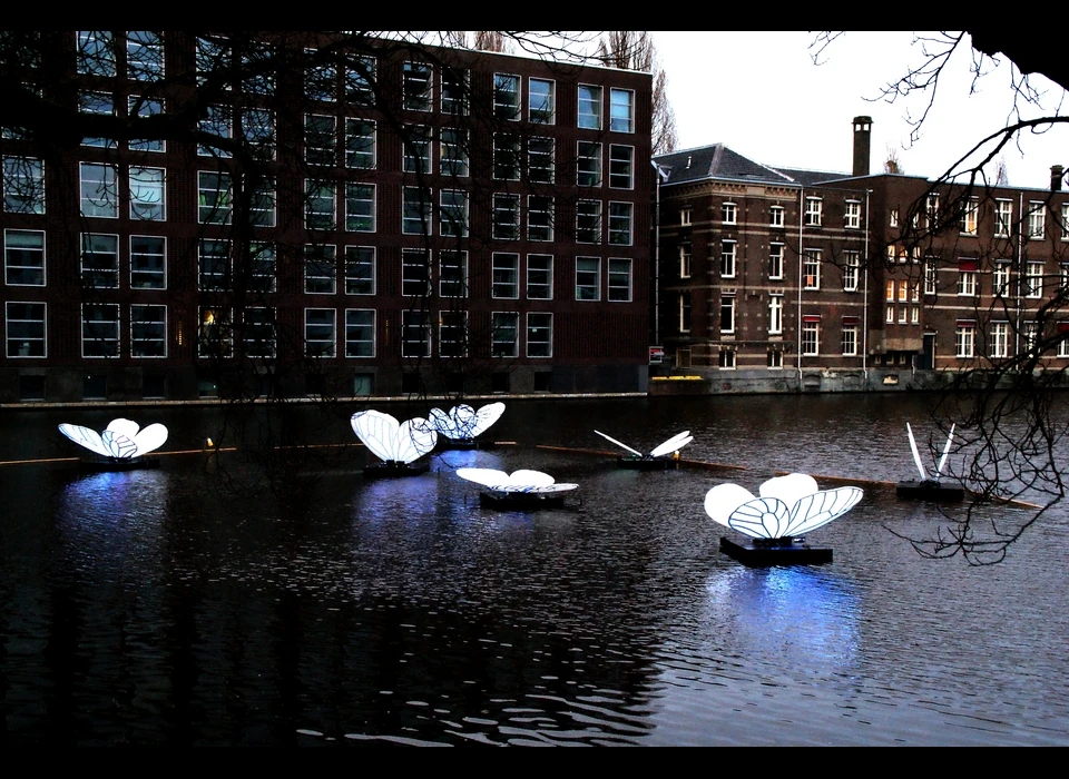 Zeven reusachtige vlinders zijn neergestreken op het 
					  wateroppervlak van de gracht. Een rimpeling veroorzaakt door een langsvarende boot doen de vlinders niet alleen op en neer deinen 
					  maar zorgen ervoor dat de vleugels van de vlinders blauw oplichten in de nacht. Op het eerste aanblik toont het kunstwerk een 
					  vredig, bijna magisch tafereel. Maar dat iets teders als een vlinder een ook een enorme kracht kan bezitten is wat kunstenaar 
					  Masamichi Shimada wil laten zien met Butterfly Effect verwijsend naar een lezing uit 1961 van de Amerikaanse wiskundige en meteoroloog 
					  Edward Lorenz getiteld, 'Predictability, does the Flap of a Butterfly's Wings in Brazil Set off a Tornado in Texas?'. 2019