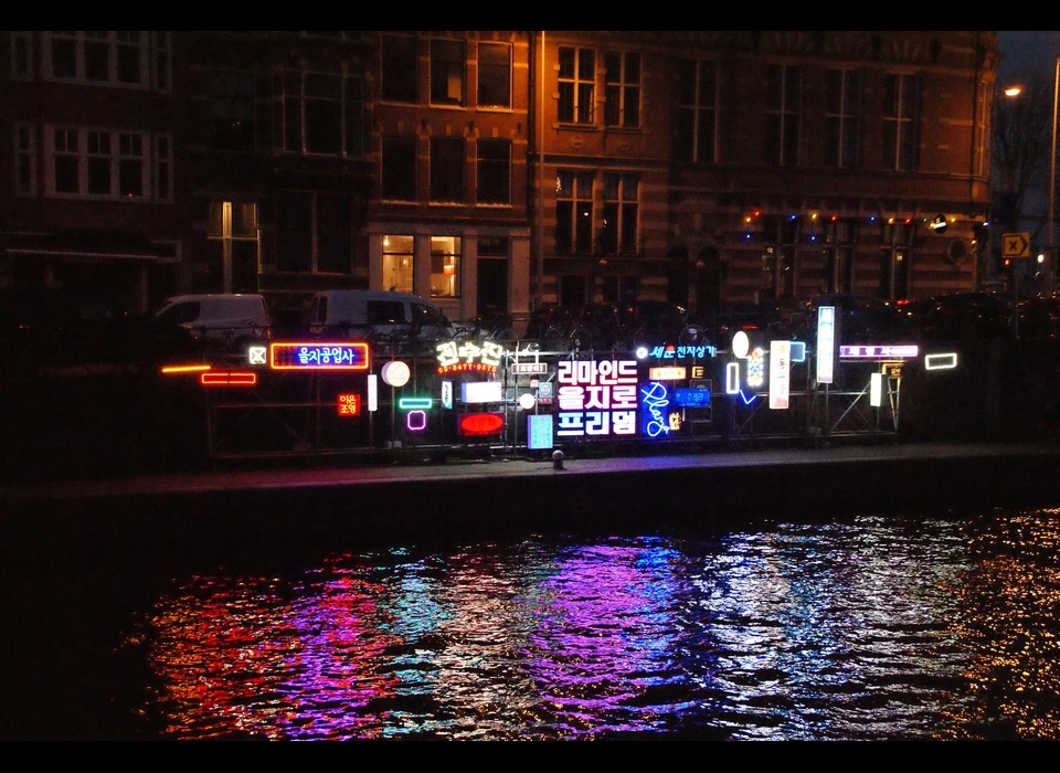 Kleurrijk flikkerende Koreaanse lichtreclames schreeuwen 
					  om aandacht in de Schippersgracht in Amsterdam. De installatie van EON SLD omvat zo’n veertig replica’s van traditionele uithangborden, 
					  neons en lichtbakken representatief voor uit een periode van de jaren vijftig tot aan het nu, en enkele nieuw ontworpen borden. De 
					  wirwar waarin deze lichtreclames zijn opgehangen aan de kade is exemplarisch voor een Koreaanse winkelstraat. 2019