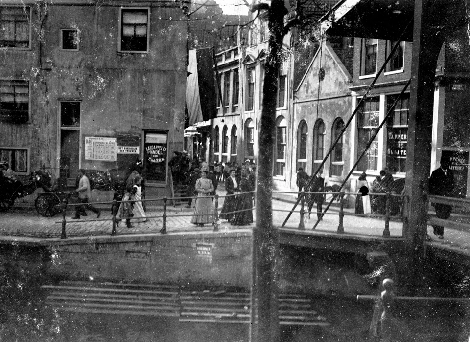 Groenburgwal 44A hoek Staalstraat 20. Het straatnaambord onder de brugboog rechts geeft aan 'Groene Burgwal'. Het plaatje in de wallekant midden onder geeft de tekst 'Brandweer, Telegraaf' en vertelt dat hier kabels door het water lopen 
					  van de brandmelders en kabels voor telegrafieverkeer. op de muur naast de etalage reclame voor het toneelstuk 'Het Koninkrijk der Vrouwen'. (1898)