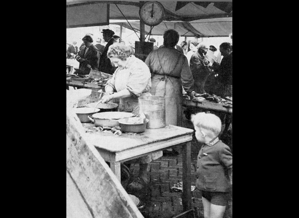 Lindengracht aalschoonmaakster bij een visboer. Het was gebruikelijk dat de 
					  gekochte aal in een pannetje werden doorgeschoven naar de schoonmaakster die voor enkele dubbeltjes de vissen onthoofde en ingewanden verwijderde. 
					  Daarna konden de spartelende dieren mee naar huis. (1955)
